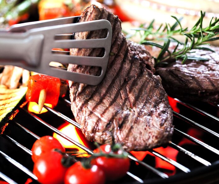 tongs holding steak on a bbq grill with rosemary and tomatoes